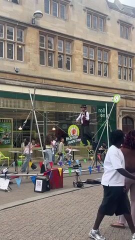 A street performer is walking across a tightrope suspended between two A-frame structures. The performer is wearing a vest, hat, and patterned pants. There are several onlookers gathered to watch, including children. In the background, there is a building with large windows and a sign for "It's British Sushi Time". Colorful flags and a speaker are set up on the ground.