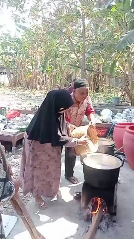 A man and a woman are in an outdoor setting, with trees and foliage visible in the background. The man, wearing a red and white patterned shirt and a black skullcap, is holding a woven basket filled with light-colored material, possibly rice. He is pouring the contents of the basket into a large black pot. The woman, wearing a black hijab and a long pink and floral patterned dress, is assisting him, holding the basket steady. The pot is positioned over a makeshift stove with burning wood, indicating they are cooking. Several other people are visible in the background, some standing and observing. There are also large red containers and stacks of bowls or plates in the vicinity, suggesting a communal cooking or event. The ground appears to be dirt or concrete. The lighting suggests it is daytime.