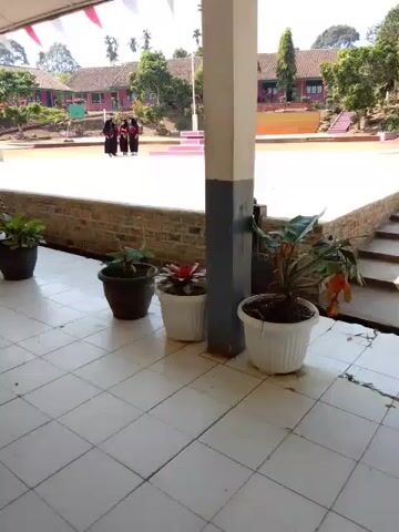 The video shows a view from under a covered walkway with tiled flooring. Several potted plants are lined up along the walkway. In the background, there is an open area, possibly a schoolyard, with a stage or elevated platform. Three individuals, appearing to be young women or girls dressed in dark clothing, are standing on the elevated platform. The environment suggests a daytime setting with clear weather, as indicated by the sunlight. The buildings in the background appear to be school structures.