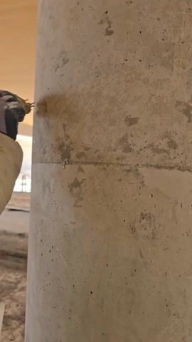 Here's a concise description of the video:

The video shows a person wearing a tan jacket and blue and gray work gloves using a small object that appears to be a brush or sponge to rub a gray concrete pillar. The pillar is textured and has visible seams. The environment is an indoor construction site. A loud noise can be heard in the background.