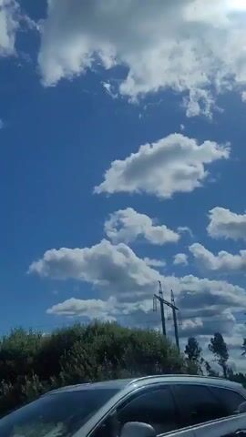 The video pans across a bright blue sky with scattered white clouds. In the foreground, a green grassy field is visible, with trees and a power line tower. The sun is shining brightly in the upper right corner, with lens flare. The camera is in motion, possibly from a vehicle.