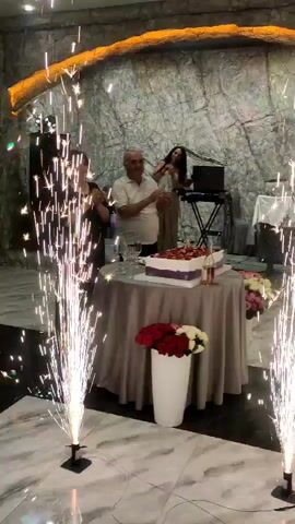 This video shows an indoor event, possibly a celebration or reception. In the foreground, two tall, thin, vertical sparkler fountains are active, shooting sparks upwards. In the mid-ground, a round table draped with a grey tablecloth is set up. On the table, there is a white cake with purple ribbon trim and a red fruit topping. Next to the cake, there appear to be champagne glasses and a bottle. In front of the table, a tall white vase holds a bouquet of red and white flowers. Behind the table, a man in a light-colored shirt is clapping. To his right, a woman in a light-colored dress stands near a DJ setup with a laptop and speakers. The background features a textured, grey stone wall with an illuminated orange archway at the top. The floor is marble.
