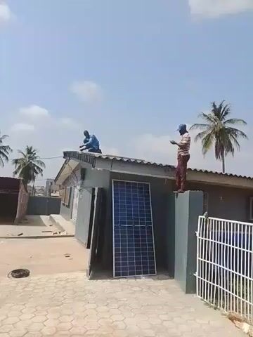 Two men are installing solar panels on the roof of a one-story building. One man is kneeling on the roof, appearing to be working on the roof itself, while the other is standing on the edge of the roof, facing forward. Several solar panels are leaning against the wall of the building. The building is gray and has windows and a corrugated metal roof. A small white gate is visible in the right corner. The ground is paved with stone bricks. Palm trees are visible in the background against a blue sky with some clouds.