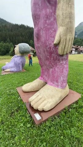 This media file features an outdoor art installation with two large, stylized human sculptures in a grassy field. The foreground is dominated by the immense feet and lower legs of a standing sculpture, clad in vibrant purple trousers. Its skin is a sandy beige, and its toes are clearly defined. Behind it, to the left, a smaller, recumbent sculpture is visible. This one is entirely purple, with a large, grey, rounded head. Its eyes are closed, and it appears to be resting or sleeping.

In the middle ground, a person wearing a red jacket and blue jeans stands between the two sculptures, providing a sense of scale to the artwork. The background shows a lush, green forest covering rolling hills, with a few buildings nestled amongst the trees. Beyond the forest, majestic mountains rise, their peaks partially obscured by the overcast sky. The weather appears to be cloudy, with a diffused light suggesting it might be daytime, possibly late morning or afternoon. The overall atmosphere is serene and artistic, with the sculptures adding a whimsical and monumental touch to the natural landscape. No visible text is present in the media file.