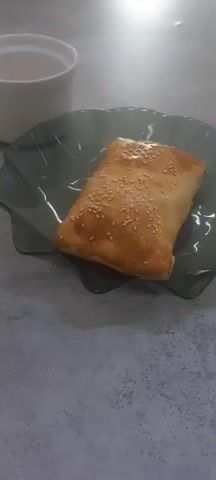 A baked pastry topped with sesame seeds sits on a dark, wavy glass plate. To the left of the plate are two white cups, one filled with a light brown liquid, presumably coffee or tea, and the other empty. The items are placed on a textured, gray surface. The scene is lit, and the camera angle is from above, looking down at the items.