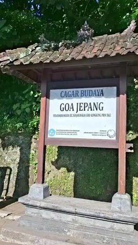 The video opens with a sign that reads "CAGAR BUDAYA GOA JEPANG" in large, bold letters. Beneath this, smaller text indicates the location: "DESA BANGLANGKAN, KEC. BANGLANGKAN, KAB. KLUNGKUNG, PROV. BALI". The sign is mounted on wooden posts in front of a moss-covered stone structure with a tiled roof. To the right of the sign, the entrance to a cave, also covered in moss, is visible. The ground in front of the sign and cave is paved with grey interlocking stones. The camera then pans to the right, revealing a tree growing alongside the paved area, with a green wall covered in vegetation behind it. Further to the right, another sign with orange and white text is partially visible. The overall environment appears to be outdoors, with abundant greenery suggesting a tropical or sub-tropical setting. The lighting suggests it is daytime. There are no people visible in the video.