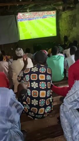 The video shows a group of people watching a soccer match on a large projector screen in what appears to be an outdoor or open-air setting. The viewers are seated on the ground, facing the screen. The lighting is dim, with the brightest light emanating from the screen itself. The people are wearing casual clothing, including head coverings and patterned shirts. The environment seems rustic, with exposed wooden beams visible overhead and dirt or packed earth on the ground. The focus is on the shared experience of watching the game, with the viewers' attention directed towards the screen.