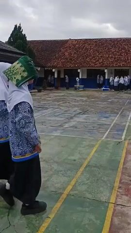 The video shows several young girls wearing school uniforms, which include white hijabs, patterned blue and white tops, and long black skirts, standing and walking around an outdoor schoolyard. One girl has a green object, possibly a backpack or bag, on her head. The schoolyard is marked with yellow lines and surrounded by school buildings with red tile roofs and blue and white trim. Other students can be seen in the background. The sky is overcast, suggesting a cloudy day. The video seems to capture a moment of students socializing during school hours.