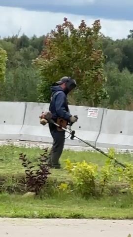 A person wearing a protective suit and helmet is using a weed trimmer to cut grass. The person is standing in a grassy area with bushes and trees in the background. A concrete barrier is visible behind the person. The sky is overcast.
