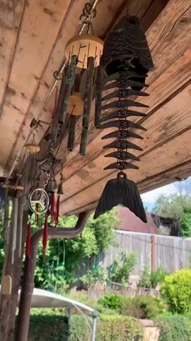 The video shows a close-up, low-angle shot of several wind chimes hanging from a wooden ceiling or awning. The wind chimes are made of various materials, including metal tubes, bells, and decorative elements such as a fish skeleton with a tail fin. The chimes are varied in design and length. Some have red tassels hanging from them. The ceiling is made of weathered wooden planks, and metal fixtures are visible for hanging the chimes. In the background, there is greenery, including bushes and trees, and a wooden fence. The lighting suggests it is daytime. The sound of the wind chimes tinkling is audible.