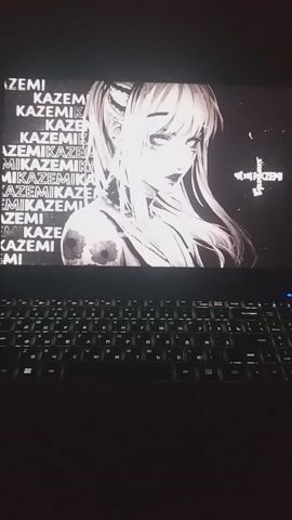 The video displays a laptop screen and part of its keyboard. The screen shows a black and white image of an anime-style character. The character has long, flowing, light-colored hair and is looking upwards and slightly to the left. A floral tattoo is visible on her left shoulder/upper arm. Below the character, the word "KAZEMIKA" is repeated multiple times in a stylized font. At the very top of the screen, a small cross-like symbol is present.

To the right and below the screen, a dark-colored keyboard with backlit keys is visible. The keys glow with a white light, illuminating the characters on them. Some of the visible letters appear to be Cyrillic, including 'И', 'Д', 'В', 'А', 'М', 'Р', 'О', 'Л', 'Ь', 'Ж', 'Э', 'Я', 'Ч', 'С', 'Ю', 'Б', 'Г', 'Ш', 'Щ', 'З', 'Х', 'Ъ', 'Ф', 'Ы', 'П', 'Р', 'О', 'Л', 'Д', 'Ж', 'Э', 'Й', 'Ц', 'У', 'К', 'Е', 'Н', 'Г', 'Ш', 'Щ', 'З', 'Х', 'Ъ'.

The camera slowly pans from the upper portion of the screen, down to the keyboard, and then back up to the screen, providing a closer look at both elements. The overall lighting in the environment appears dim, highlighting the screen's image and the keyboard's backlighting.