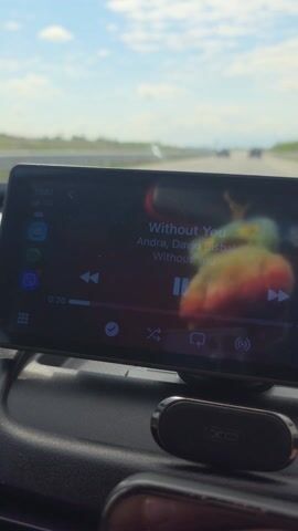 A camera is mounted on the dashboard of a car. The car is driving on a highway. The sky is blue with some clouds. The dashboard is black. A small device is mounted on the dashboard, displaying song information for "Without You" by Andra, David Bisbal. The speedometer needle indicates the car is traveling under 100 kilometers per hour.