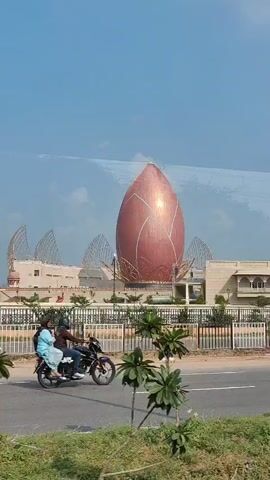 A video captures a large, pink, bud-shaped structure resembling a lotus flower from a moving vehicle. The structure is ornate with golden lines and is surrounded by architectural elements and greenery. Several vehicles, including cars and a motorcycle with two passengers, are visible on a road in the foreground, suggesting the footage is taken from a street or highway. The sky is clear and blue, indicating daytime.