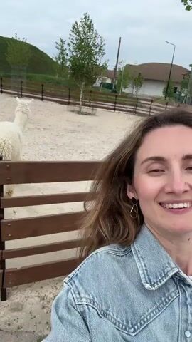 The video shows a woman taking a selfie in front of a wooden fence, with a white alpaca in the background. She is smiling and wearing a denim jacket. Another person with blonde hair and a red shirt is also visible in the background, holding a phone. The environment appears to be a farm or zoo-like setting, with a grassy hill and trees in the distance. The ground is sandy or dirt-covered.