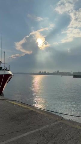 The video presents a scenic shot of a body of water, possibly a bay or river, with a cityscape visible on the distant horizon. The sky is partially covered by clouds, with the sun attempting to break through, casting rays of light onto the water's surface and creating a shimmering effect. A portion of a boat or ship is visible in the bottom left corner of the frame, docked at a concrete pier or quay. The water is calm, with gentle ripples reflecting the sunlight. The overall atmosphere is serene and somewhat dramatic due to the contrasting light and shadows.