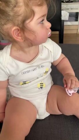 A toddler sits on a gray couch, sucking on a pacifier. They are wearing a white onesie that says "EVERYDAY I WANT TO BEE..." and has bees printed on it. Their socks are white. In the background, a TV displays a cartoon with a Smurf character. A children's slide and other toys are scattered on a rug in the living room.