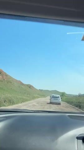 This video depicts a point-of-view shot from inside a car traveling on a dusty, rural road. The scene is set in a hilly, green landscape under a clear blue sky, suggesting a bright, sunny day. Several silver cars are visible ahead on the road, indicating a convoy or a traffic situation. The car in the foreground is following a silver hatchback.

The road is unpaved and appears to be in fair condition, with visible ruts and loose gravel. The surrounding terrain is characterized by rolling hills covered in dry grass, with some patches of darker green vegetation. In the distance, more hills recede into the horizon. The overall atmosphere appears to be one of peaceful travel through a natural, undeveloped area.

A yellow sticker with the number "83" is visible on the upper right corner of the windshield. The license plate of the silver hatchback in front is partially visible and appears to read "E 929 ET 102". The location is Drokino, Russia. The time of day is clearly daytime, likely midday due to the bright sunlight and clear skies. There are no people visible outside the cars. The activity is simply driving along the road.