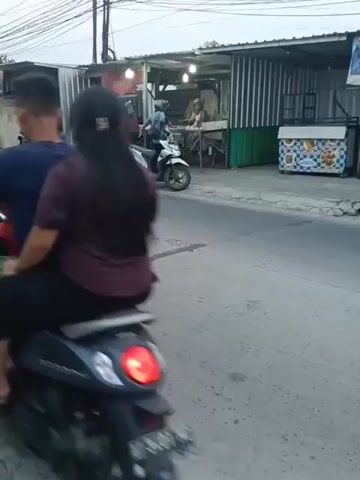 The video shows a street scene with several motorcycles and a yellow car. There are small food stalls or shops lining the side of the road. A white motorcycle with two people on it is in the foreground, moving from left to right. To the left of the white motorcycle, a yellow car is also moving from left to right. Further in the background, other motorcycles and people can be seen. The environment appears to be an urban or suburban setting with overhead power lines. The overall atmosphere is that of daily life on a street.