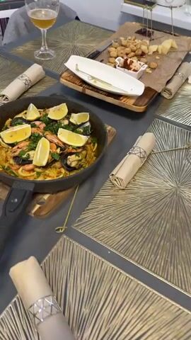 The video shows a close-up, slightly angled view of a paella dish in a black, cast-iron skillet. The paella is garnished with lemon wedges and fresh herbs, and appears to contain shrimp and mussels. The skillet is resting on a wooden cutting board. In the background, a table is set with placemats, napkins, and a glass of beer. There is also a wooden board with cheese, nuts, and some red dip. A lit candle in a glass holder is visible in the foreground. The scene appears to be a meal setting, possibly a dinner.