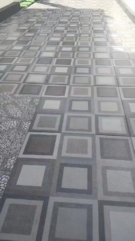 A video pans across a tiled walkway leading to a gate. The walkway is made of square tiles with a geometric pattern of concentric squares in shades of gray. To the left, a white wall is visible, and to the right, another white wall defines the path. In the background, a dark metal gate with vertical bars stands closed. Behind the gate, lush green foliage, including large banana leaves, is visible. Overhead, power lines crisscross the clear blue sky. The scene appears to be an outdoor residential area.