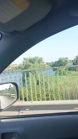 This media file is a video recording taken from inside a vehicle, likely a car, as it travels across a bridge. The perspective is from the passenger side, looking out the window.

**Subjects:** The primary subject is the view from the moving vehicle. This includes a body of water, possibly a river or lake, stretching out in the distance. On the far bank, there are trees and some buildings, suggesting a populated area. A metal railing, painted with vertical bars of green and yellow, runs along the side of the bridge, partially obscuring the view of the water and the landscape beyond. The interior of the car, including a side mirror and the car door frame, is also visible in the foreground.

**Activity or Event:** The video captures the experience of traveling across a bridge. The slight shake and the changing perspective of the side mirror indicate the vehicle is in motion. The scene is one of passing over a watery expanse.

**Notable Details:**
*   **Time of Day:** The bright, clear sky suggests it is daytime, likely late morning or afternoon, given the lighting.
*   **Weather:** The clear sky indicates fair weather.
*   **Location Cues:** The presence of a bridge over a body of water, with trees and buildings on the far bank, is consistent with a riverside or lakeside town. The location context provided is Maiaky, Ukraine, which aligns with a landscape that could feature such a scene.
*   **Emotions:** The footage evokes a sense of travel and perhaps peaceful observation of the passing scenery. There are no people clearly visible to infer emotions from.

**Visible Text:** No discernible text is visible in the video.