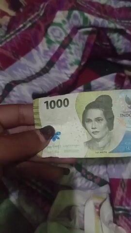 A person's hand holds a 1000 Indonesian Rupiah banknote. The banknote features a portrait of a woman, the number "1000", and the Garuda Pancasila emblem. The banknote is placed on a colorful, patterned fabric. The background is out of focus and shows the same patterned fabric.