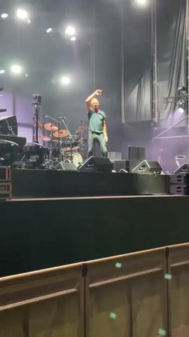 A man wearing a blue t-shirt and dark pants is on a stage, facing left and looking down with a slight smile. He is holding a microphone in his right hand and gesturing with his left hand, which is raised in the air. Behind him, a drum set and various audio equipment are visible. The stage is lit with bright spotlights. In the foreground, a blurred hand is visible, suggesting the video is being recorded by someone in the audience.