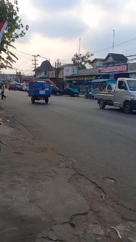 A street scene at dusk shows traffic on a paved road. A blue utility vehicle with a trailer drives away from the camera on the left side of the road. A white pickup truck, a blue pickup truck, and several cars are visible on the road. A motorcycle with two riders is passing the white pickup truck. A motorcycle with one rider is on the right side of the road. Buildings line the right side of the road, some with awnings displaying signs. The sky is cloudy.