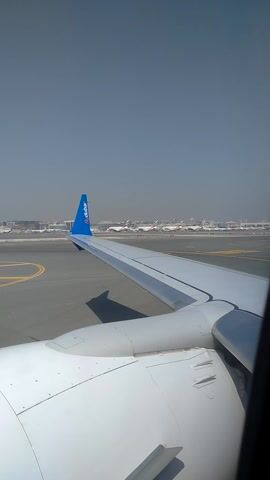 This video appears to be filmed from the window of an airplane as it taxis on a runway. The perspective is looking out over the wing and the engine of the aircraft. The wing is white with a blue wingtip that has the "flydubai" logo on it. The runway is asphalt with yellow markings. In the distance, there are several other airplanes lined up, suggesting an airport. The sky is a hazy blue, indicating possibly a bright but dusty or polluted atmosphere, or it could be early morning or late afternoon light. The overall scene is one of travel and anticipation, likely at the start of a journey. There is no visible indication of people within the frame, only the interior of the aircraft's cabin edge at the top right corner of the image. The time of day is not explicitly stated but the lighting suggests it is daytime. The location is given as Ilidža, Bosnia and Herzegovina, which is not directly evident from the visual cues in the image, but it is possible this is the departure airport.