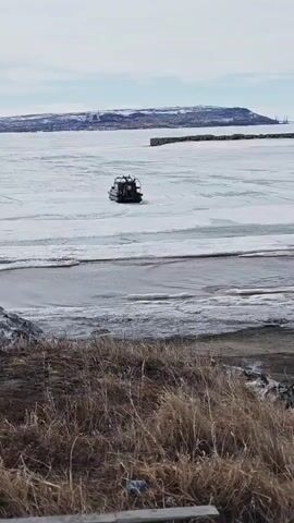 A hovercraft traverses a frozen expanse of water. In the foreground, dry grasses cover the bank. Further out, a body of water partially covered in ice extends towards a distant land mass. The sky is overcast. The hovercraft, dark in color, moves smoothly across the ice.  A small stretch of open water is visible between the bank and the frozen area. The land mass in the distance includes what appears to be a small town and some industrial structures.