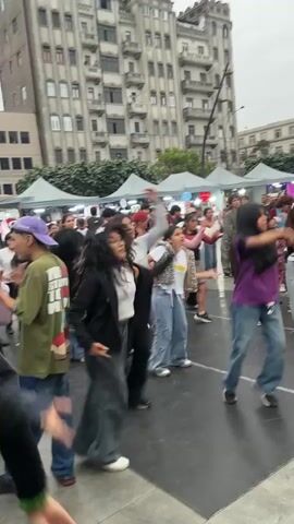 A crowd of young people are dancing in a square with a large building in the background. Most of the dancers are wearing baggy jeans. White tents are visible behind the crowd, suggesting a market or event is taking place. The weather looks overcast.