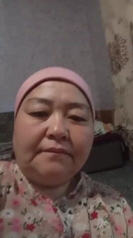 This is a selfie-style video of a middle-aged woman wearing a light pink headscarf and a floral patterned shirt. She is looking directly at the camera. The background is out of focus, but appears to be an indoor setting. To her left, a dark wardrobe or cabinet is visible. To her right, a couch with a patterned fabric is partially visible. There is a textured, light-colored wall behind her. The lighting suggests it is daytime. The woman's expression shifts from a neutral gaze to a smile. No text is visible in the media file. The location context is Chon-Kara, Kyrgyzstan.