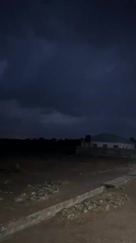 The video captures a night scene in Barako, Nigeria, dominated by a dark, stormy sky. The time is clearly evening or night, indicated by the pervasive darkness. Dark, heavy clouds fill the upper portion of the frame, occasionally illuminated by bright flashes of lightning. These lightning strikes are the primary activity, intermittently lighting up parts of the sky and briefly revealing the cloud formations.

The foreground shows a rough, earthy terrain, possibly a field or undeveloped land, with scattered small rocks and debris. A concrete curb or drainage structure runs horizontally across the lower part of the frame. In the distance, silhouetted against the slightly brighter horizon or during lightning flashes, are indistinct buildings or structures. One building with a light-colored roof and possibly white walls is visible to the right, with what appears to be a water tank on its roof or nearby. The overall impression is one of a dramatic, storm-filled night in a semi-rural or developing area. There is no visible text or human activity.