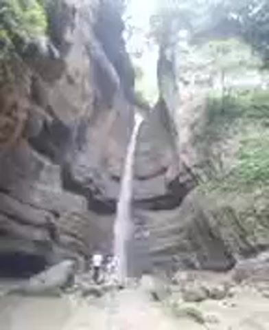 A narrow waterfall cascades down a rocky cliff face into a pool of water. Several people stand at the base of the waterfall, observing the scene. The surrounding environment is lush with green foliage, suggesting a natural, outdoor setting. The overall impression is one of natural beauty and tranquility.