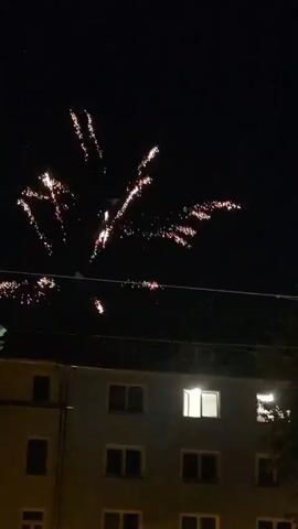 The video captures a nighttime scene with fireworks exploding in the sky above a multi-story building. The building appears to be residential, with several windows visible, some of which are illuminated. Power lines run horizontally across the frame in front of the building. The fireworks display various colors and patterns, with some bursts of white and pink, and occasional green sparks. The explosions are spread across the sky, with some appearing closer and more intense than others. The overall atmosphere is festive, suggesting a celebration or event.