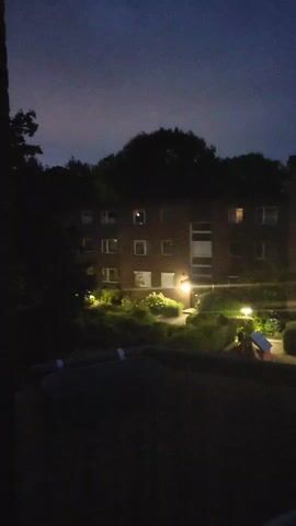 The video shows a nighttime view of an apartment building with trees surrounding it. There are lights on in some of the windows, and landscape lighting illuminates the bushes and plants in front of the building. A small playground structure with red and blue elements is visible in the lower right portion of the frame. The sky is dark with a few clouds. The camera appears to be positioned on a balcony or from a window looking out at the scene.