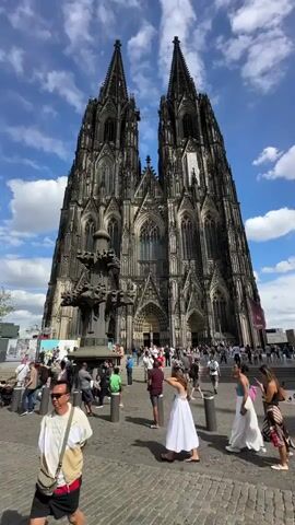 The video shows a wide-angle view of Cologne Cathedral in Germany under a partly cloudy sky. The massive Gothic cathedral dominates the frame with its two spires. In the foreground, a cobblestone plaza is filled with people milling about. A dark, ornate fountain structure is visible to the left of the cathedral. Several individuals are walking and standing, some appear to be tourists. The overall atmosphere seems to be that of a busy public space on a bright day.