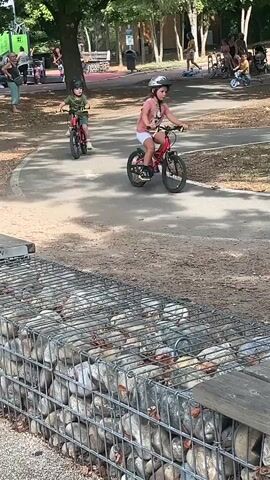 Two children are riding bicycles on a paved path in a park. The child on the right is wearing a pink shirt and white shorts, and the child on the left is wearing a green shirt and shorts. Both children are wearing helmets. The path is surrounded by grass and trees. There are other people and children in the background, some sitting on a bench and others riding bicycles. The video also shows a wire mesh fence filled with rocks in the foreground.