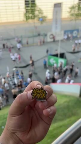 This media file captures a lively outdoor scene with many people gathered. In the foreground, a hand holds up a small, yellow and black pin. The pin displays text that appears to read "MARCCA KOPP" above "DÜSSELDORF 2021".

The background reveals a large crowd of people, suggesting an event or festival is taking place. Many individuals are standing and milling about, while others are seated on a grassy slope, enjoying the atmosphere. Several red tents are visible, possibly serving as food or merchandise stalls. Some people are dressed in casual summer attire, and the overall mood seems relaxed and energetic.

The scene is set outdoors during daylight hours, likely late afternoon or early evening given the angle of the sun and the warm lighting. Trees and a modern building with a distinctive slatted facade frame the background, hinting at an urban or stadium-like environment. The presence of the "DÜSSELDORF 2021" text on the pin strongly suggests the location is Dusseldorf, Germany, and the event took place in 2021. The activity appears to be a social gathering, possibly a concert, festival, or sporting event, with attendees enjoying the warm weather and communal experience.