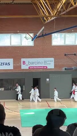 The video captures an indoor sports gymnasium with a wooden floor, red brick walls, and a wooden beam ceiling structure. Two basketball hoops are visible overhead. On the floor, several individuals, primarily children and some adults, are present. Most wear white martial arts uniforms (doboks); some also wear red and blue protective vests and helmets. On the left, an adult in a white dobok and dark belt walks right, followed by three children in doboks. In the center, a child in a white dobok performs a martial arts pose, raising their right fist. On the right, four individuals (three children, one adult-sized) in doboks with protective gear walk left. In the background, two banners are affixed to the wall, one displaying "ZFAS" and another "B_barcino" with a web address. The tops of several spectators' heads are visible in the blurred foreground, watching the activity.