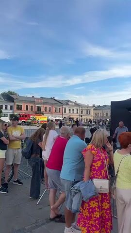 The video shows an outdoor event in front of a large, ornate building, likely a town hall or historical structure, with a tall tower. A crowd of people, mostly adults and some children, are seated on chairs facing a stage or performance area. A large screen displays colorful graphics with text in Polish, likely announcing the event. There are tents and equipment set up on the stage area, suggesting a concert or public performance. The sky is partly cloudy and the atmosphere appears festive. Some people are standing and watching the event, while others are seated. The camera pans across the crowd and the building, capturing the scene from different angles.