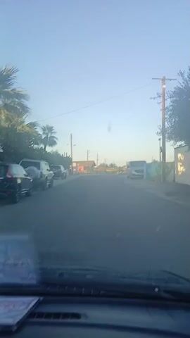 The video captures a drive from the perspective of a vehicle's interior, looking out through the windshield. The dashboard and wipers are visible at the bottom of the frame. The setting is a street in Nisou, Cyprus, under a clear, bright blue sky, indicating daytime.

The car initiates a right turn at the beginning of the video. The road is paved with asphalt. Along the sides of the street, there are power poles with overhead lines, various trees including palm trees, and scattered single and two-story buildings, consistent with a residential or semi-rural area.

A white van is briefly visible parked on the right side of the road at the start of the turn (0:01). After completing the turn, a grey van is prominently parked on the right (0:03-0:04), adjacent to a wall that appears to have some painted designs or graffiti. No specific text on the wall or the van's license plate is clearly decipherable. The surroundings suggest a quiet area with houses and open plots of land. The weather is sunny and clear throughout the drive.