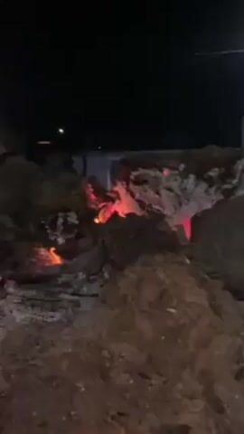The video shows a small bonfire burning at night. The embers glow orange and red. The camera then pans to show a church decorated with white lights and a blue star. There are streetlights nearby and houses across the street.