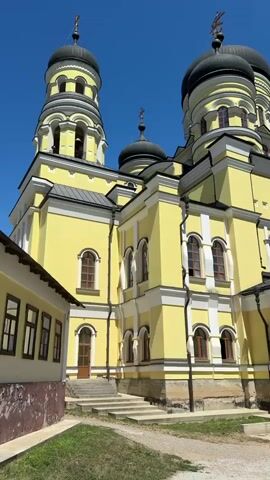 The video shows a bright yellow church with black domes against a clear blue sky. The camera pans across the side of the church, revealing multiple windows with arched tops and brown frames. There are also bell towers with similar architectural features. In the foreground, there is a pathway and some greenery. The building appears to be an Orthodox monastery or church, given the style of its domes and crosses.