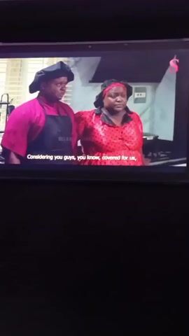 The video shows a close-up of a tablet screen displaying a scene from a show. Two women are visible, standing indoors. The woman on the left wears a yellow shirt and the woman on the right wears a black tank top. They appear to be in conversation, with the woman on the left gesturing with her hands. Subtitles are displayed at the bottom of the screen. The scene transitions briefly to show a third woman in a red and white polka dot top standing between the first two women, all three looking surprised. The scene then returns to the first two women.