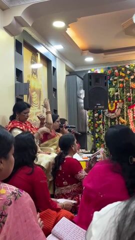 The video shows a room full of people gathered for what appears to be a religious event. Most attendees are women, many wearing red or pink traditional Indian clothing. Several women near the front appear to be leading the ceremony, one holding a microphone and singing. A large speaker is set up to the right. Behind them is an elaborate floral display featuring images of Hindu deities. On the left is a wall with an image of Buddha. The ceiling is brightly lit with recessed lighting and a decorative chandelier.