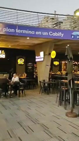 The video shows an outdoor dining area under an overpass in an urban environment. Several people are seated at high-top tables with black stools. Some are conversing, while others are looking at their phones. Overhead signs advertise Cabify and mention "El mejor plan". A KFC restaurant is visible in the background. Buildings line the background. The ground is paved with gray stones. There are several black umbrellas providing shade. The lighting suggests it is late afternoon or early evening.