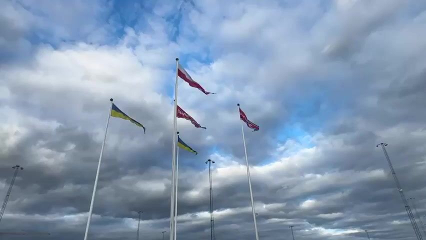The video displays a low-angle view of several flags flying on poles against a cloudy sky. From left to right, there are two Ukrainian flags, one Polish flag, and two red flags with a crest. The flags are positioned in front of a large, modern building with a tall tower on the left side of the frame. In the background, a parking lot is visible, filled with numerous cars and trucks. The sky is a mix of dark and light gray clouds with patches of blue sky. The overall scene suggests an outdoor location, possibly a public space or an industrial area.