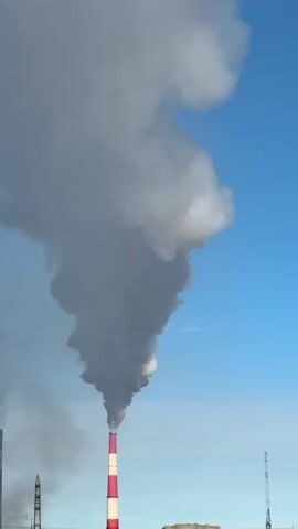 A tall, red and white striped smokestack emits a thick plume of grey smoke against a clear blue sky. The smoke billows upwards and outwards, forming a large cloud that dominates the frame. In the background, to the left of the smokestack, another shorter, dark smokestack is partially visible, also emitting a thinner stream of smoke. Further to the right, a thin, tall metal structure, possibly a transmission tower, stands against the sky. The lower portion of the image shows a faint glimpse of industrial buildings or structures. The overall scene depicts an industrial area with active emissions.