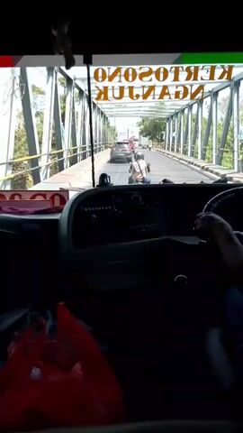 The video is filmed from the perspective of someone inside a bus, looking forward through the windshield. The bus is on a bridge, indicated by the metal structure spanning overhead with the words "KERTOSONO MADIUN" visible, though upside down and reversed. A driver's hand is visible on the steering wheel. Ahead, traffic is moving across the bridge, including several motorcycles and a car. The environment outside the bridge appears to be lush with trees and greenery. In the foreground inside the bus, a red bag is visible on the left.